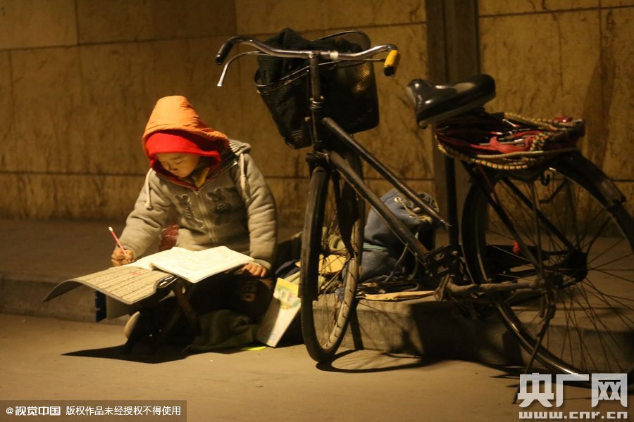 鄭州中原路京廣鐵路大橋下涵洞的馬路牙子邊，一位戴著紅色辣椒帽的小男孩趴在一個小馬扎上，上面鋪著一張車墊，在微弱的燈光下寫作業(yè)